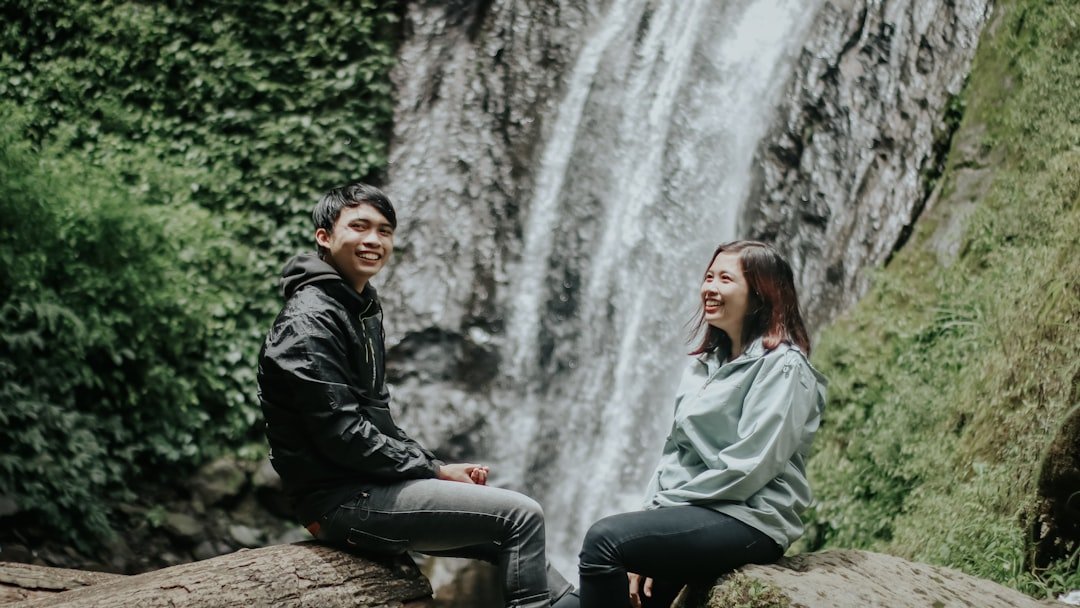 man in black leather jacket sitting on rock near waterfalls during daytime